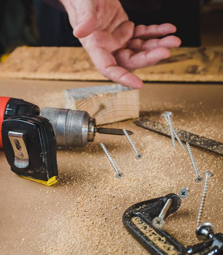 Plan de travail d’un atelier avec outils de menuiserie, vis, perceuse, serre-joint et main d’un artisan, d'Ovan