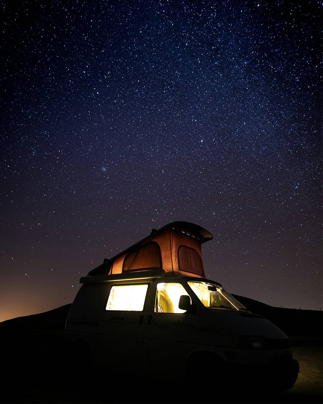 Van aménagé avec toit relevable éclairé sous un ciel étoilé, idéal pour une nuit en pleine nature.