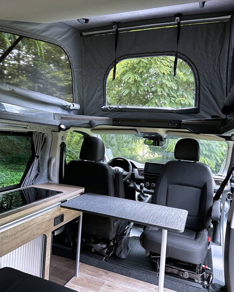 Intérieur d’un van aménagé avec toit relevable, kitchenette en bois, table pliante et vue sur l'extérieur.