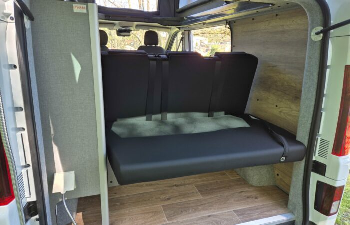 L'image montre l'intérieur d'un camping-car avec une banquette noire rabattue, des ceintures de sécurité, un plancher imitation bois et un panneau gris clair avec des prises électriques à gauche. La lumière du soleil pénètre par les fenêtres.