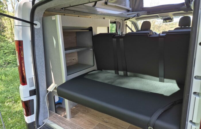 L'intérieur d'un camping-car avec une banquette, deux étagères, un plancher imitation bois et de grandes fenêtres laissant entrer la lumière naturelle ; la porte du camping-car est ouverte sur un espace extérieur herbeux.