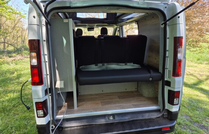 L'image montre l'arrière ouvert d'un camping-car blanc garé sur l'herbe, révélant un intérieur épuré avec un plancher imitation bois, une banquette noire et des panneaux clairs. Des arbres et de la verdure sont visibles en arrière-plan.