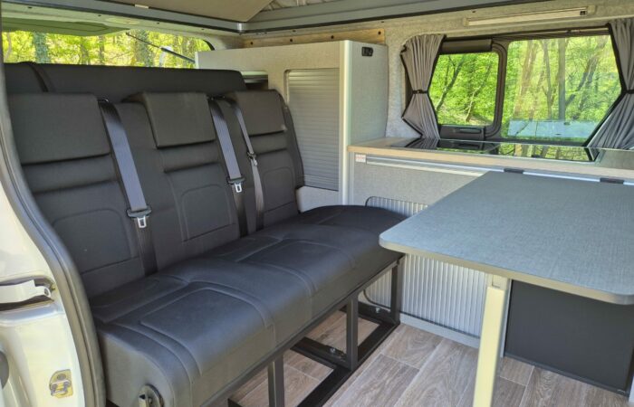 Intérieur d'un camping-car comprenant une banquette noire avec ceintures de sécurité, une table grise, un plancher imitation bois et de grandes fenêtres avec rideaux, avec des arbres verts visibles à l'extérieur.