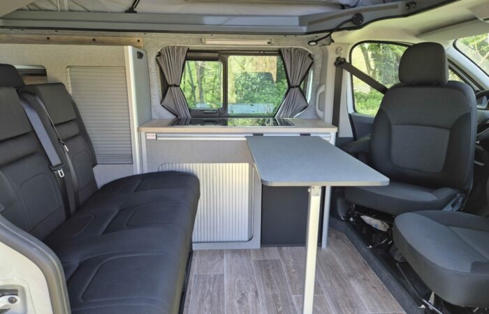 Intérieur d'un camping-car avec sièges noirs, table pliante, kitchenette compacte, petits rideaux aux fenêtres et parquet aspect bois, avec des arbres visibles à l'extérieur à travers les fenêtres.