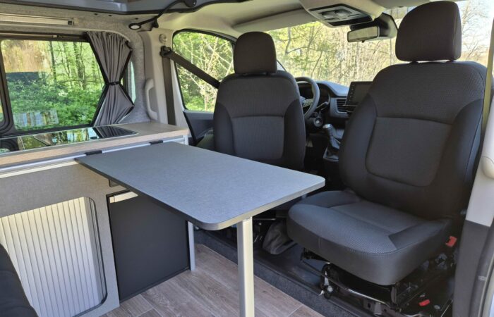 Intérieur d'un camping-car avec deux sièges avant noirs, une table grise, un plancher imitation bois, une kitchenette compacte avec un évier et de grandes fenêtres montrant des arbres verts à l'extérieur.