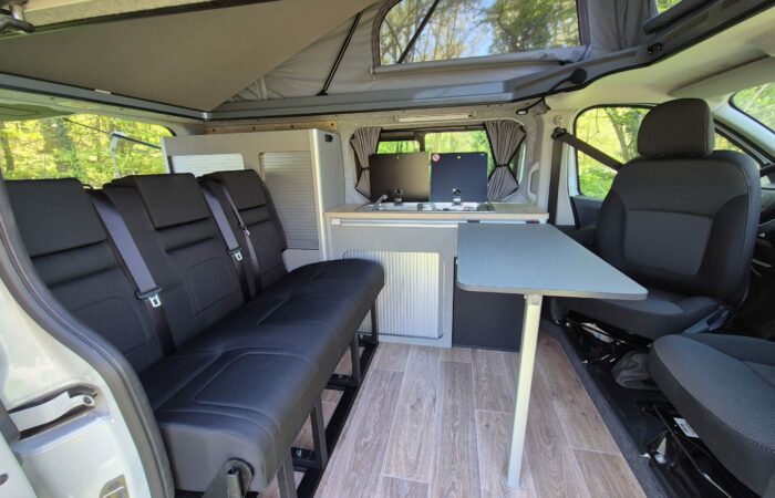 Intérieur d'un camping-car moderne avec sièges noirs, table pliante, kitchenette avec cuisinière et évier, plancher en bois clair et toit relevable. La lumière du soleil pénètre à travers les fenêtres, créant un espace chaleureux et organisé.