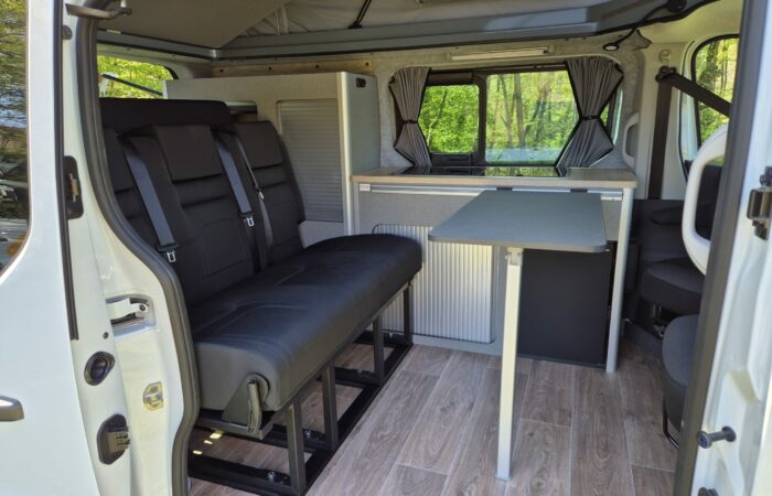 L'intérieur d'un camping-car comprenant des banquettes noires, une table, un parquet en bois clair et une kitchenette compacte avec des armoires et de petites fenêtres avec rideaux, entourée de verdure visible à l'extérieur.