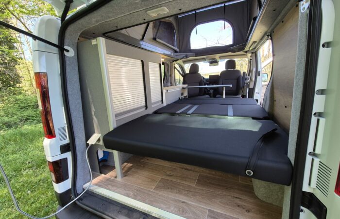 Vue de l'arrière d'un camping-car montrant une plate-forme de lit pliée, des parois intérieures grises, un plancher en bois, deux sièges avant et de grandes fenêtres avec des arbres visibles à l'extérieur.