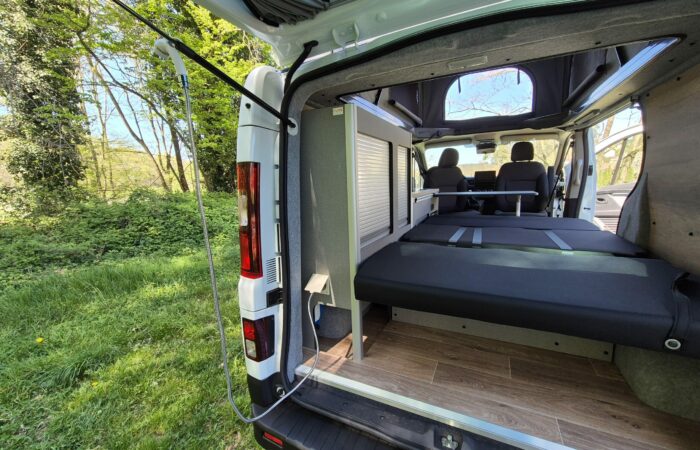 Vue depuis les portes arrière ouvertes d'un camping-car, montrant un lit pliant, des compartiments de rangement, un plancher imitation bois et le camping-car garé dans une zone herbeuse et boisée par une journée ensoleillée.