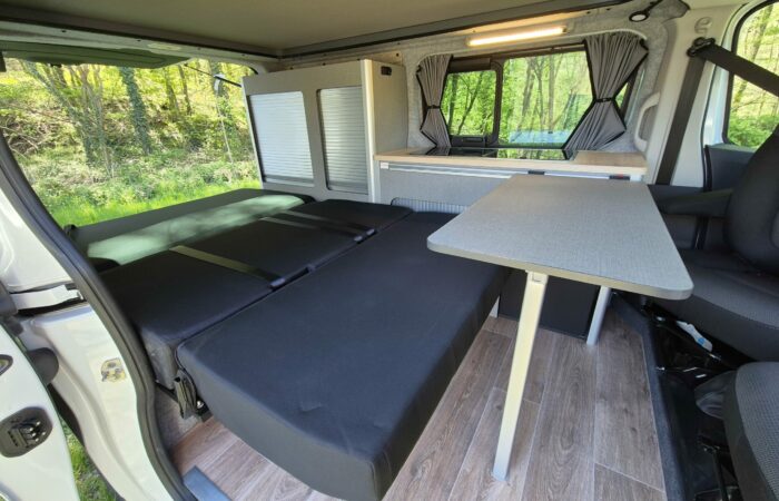 Intérieur d'un camping-car moderne avec un lit noir déplié, une table grise, un parquet imitation bois et des fenêtres avec rideaux, garé dans un espace vert et boisé.