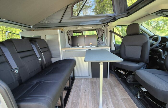 Intérieur d'un camping-car avec sièges noirs, table pliante, parquet en bois clair, coin cuisine compact et toit ouvrant ouvrant laissant entrer la lumière naturelle de la verdure environnante.