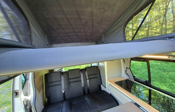 Intérieur d'un camping-car avec un toit relevable surélevé, trois sièges noirs, une petite kitchenette avec un évier et une cuisinière, et la lumière du soleil traversant les fenêtres latérales, offrant une vue sur l'herbe verte à l'extérieur.