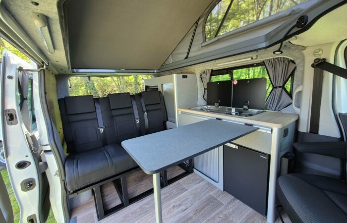 Intérieur d'un camping-car moderne avec table pliante, banquette noire, kitchenette compacte, réfrigérateur et fenêtres avec rideaux. La lumière du soleil pénètre par la porte latérale ouverte et les fenêtres.