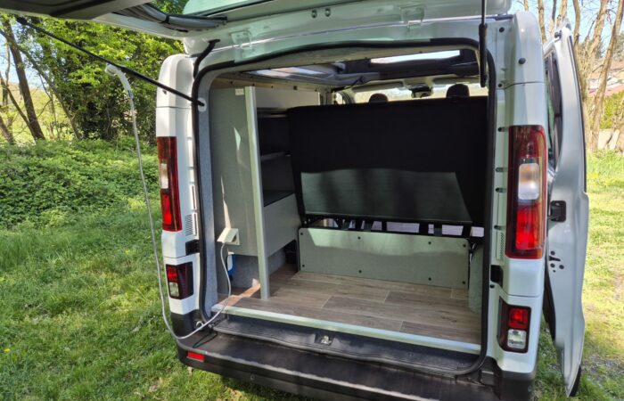 L'arrière d'un camping-car blanc avec ses portes arrière ouvertes, montrant un intérieur bien rangé avec du parquet, des étagères et une banquette, garée sur l'herbe près des arbres.