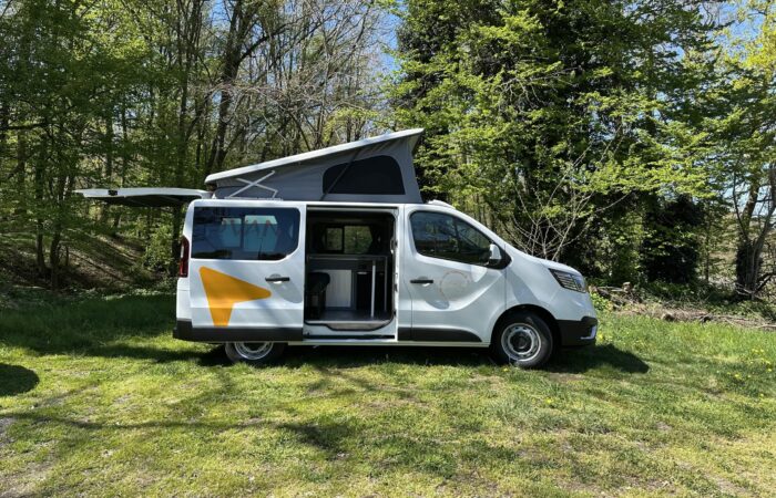 Un camping-car blanc avec toit relevable et porte arrière ouverte est garé sur l'herbe, entouré d'arbres et de soleil, révélant une cuisine compacte et un espace de rangement à l'intérieur.