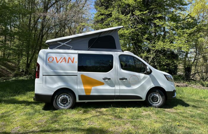 Un camping-car blanc, portant l'inscription « OVAN » et une flèche jaune sur le côté, est garé sur l'herbe sous des arbres feuillus. Son toit relevable est déployé, indiquant qu'il est prêt à camper.