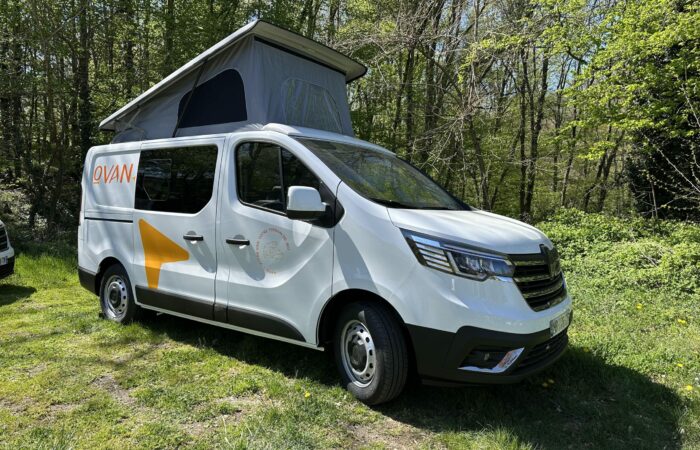 Un camping-car blanc à toit relevable est garé sur l'herbe dans une zone boisée. Le van arbore des graphismes et une marque orange sur le côté. La lumière du soleil filtre à travers les arbres verts qui l'entourent.