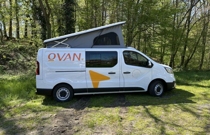 Un camping-car blanc à toit relevable est garé sur l'herbe, près des arbres. Le van arbore l'inscription « OVAN » et une flèche orange sur le flanc. La lumière du soleil filtre à travers le feuillage vert environnant.