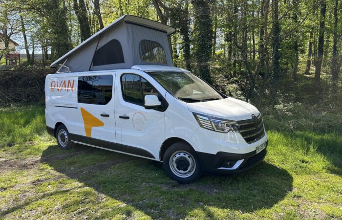 Un camping-car blanc à toit relevable est garé sur l'herbe, en bordure d'une zone boisée. La lumière du soleil filtre à travers les arbres, et le van arbore le logo « OVAN » et une flèche orange sur le flanc.