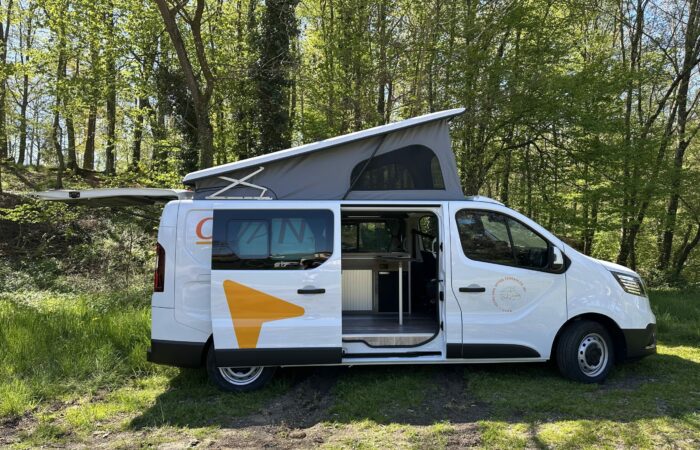Un camping-car blanc, avec sa porte coulissante et son toit relevable ouverts, est garé sur l'herbe près d'une forêt, révélant une cuisine compacte à l'intérieur. La lumière du soleil filtre à travers les arbres en arrière-plan.