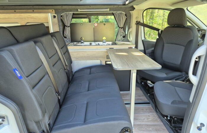 Intérieur d'un camping-car montrant deux sièges avant, une table pliante, une banquette, un plancher imitation bois et une kitchenette compacte avec un évier et une cuisinière à l'arrière, avec des rideaux sur les vitres arrière.