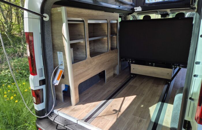 L'image montre l'intérieur d'un camping-car avec un plancher imitation bois, des étagères ouvertes intégrées, un panneau noir mat et des sièges. Les portes arrière sont ouvertes, révélant la verdure et la lumière du soleil.
