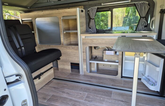 Intérieur d'un camping-car avec plancher imitation bois, banquette noire, kitchenette intégrée, petit réfrigérateur, étagères, table pliante et grandes fenêtres avec rideaux. La lumière du soleil entre à flots depuis l'extérieur.