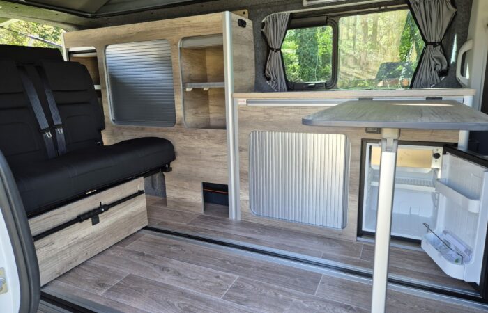 Intérieur d'un camping-car moderne avec plancher imitation bois, table pliante, banquette noire rembourrée, placards de rangement intégrés, petit réfrigérateur et fenêtres ornées de rideaux gris. La lumière naturelle pénètre à flots depuis l'extérieur.