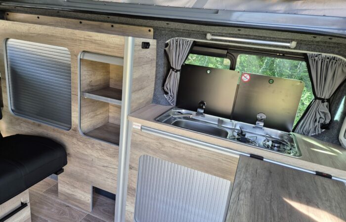 Intérieur de camping-car moderne comprenant une kitchenette compacte avec un évier et une cuisinière, des armoires lambrissées, de petites étagères et une fenêtre avec des rideaux laissant entrer la lumière naturelle.