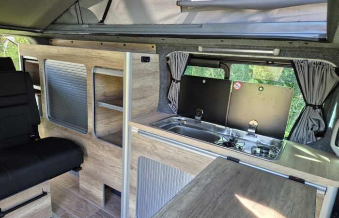 Intérieur d'un camping-car comprenant une petite cuisine avec un évier et une cuisinière, des armoires en bois pour le rangement, un siège rembourré noir et une fenêtre avec des rideaux laissant entrer la lumière naturelle.
