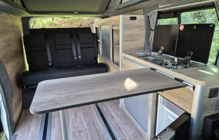 L'intérieur d'un camping-car comprend une kitchenette en bois avec évier, cuisinière et table pliante. À l'arrière-plan, des sièges passagers noirs et une fenêtre avec rideaux. La lumière naturelle pénètre par la porte ouverte.