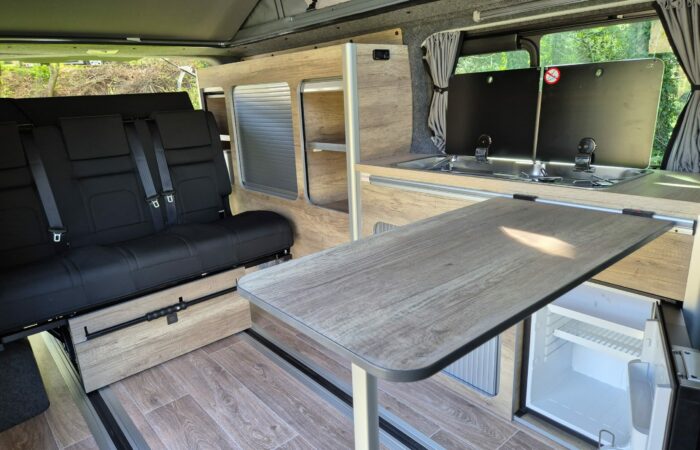 L'intérieur d'un camping-car comprend une banquette noire, une table pliante en bois, une kitchenette compacte avec évier et cuisinière, des placards et un petit réfrigérateur. La lumière du soleil filtre par une fenêtre voisine.