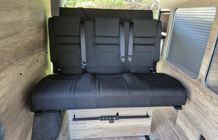 Une banquette en tissu noir avec ceintures de sécurité est installée dans une camionnette aux parois et au plancher lambrissés. Le siège est orienté vers l'avant et une fenêtre avec un volet roulant argenté fermé se trouve sur le côté droit.