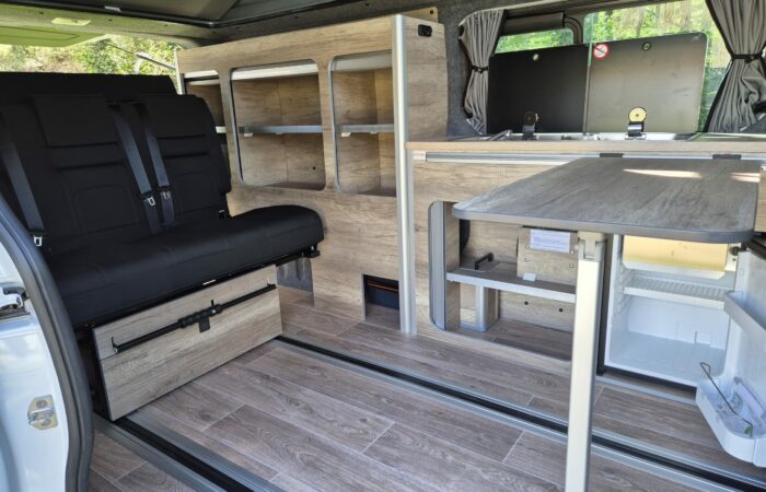 Intérieur d'un camping-car avec planchers lambrissés, une table pliante, des étagères ouvertes intégrées, un réfrigérateur et une banquette rembourrée noire avec ceintures de sécurité près des fenêtres.