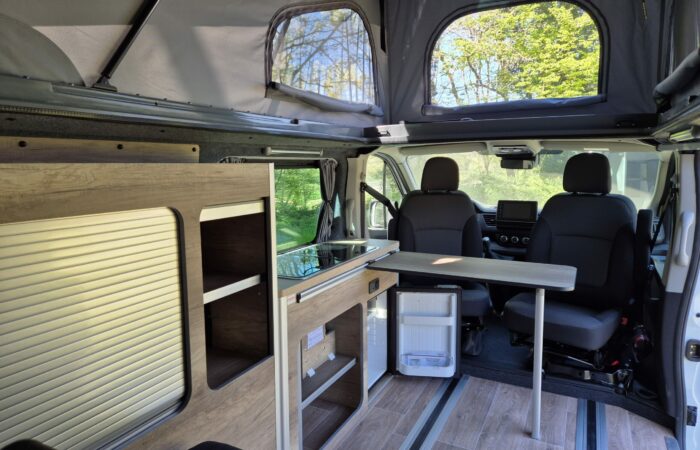 Intérieur d'un camping-car avec un toit relevable surélevé, un coin cuisine avec armoires et évier, un mini-réfrigérateur, une table et deux sièges avant ; de grandes fenêtres montrent des arbres verts et la lumière du soleil à l'extérieur.