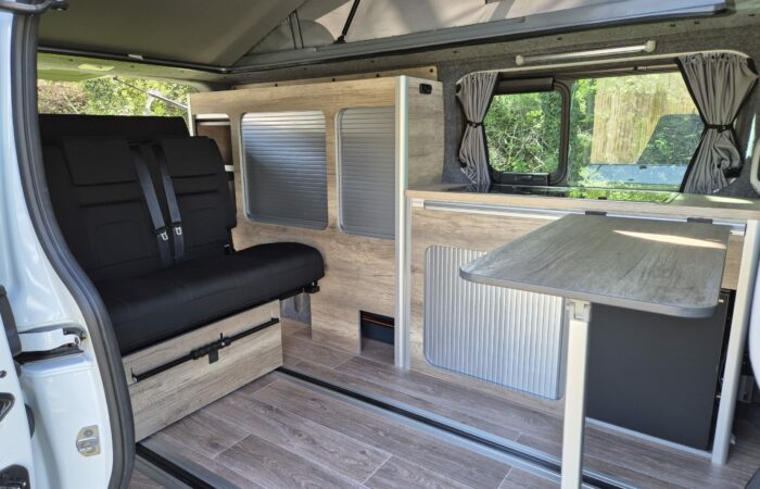L'intérieur d'un camping-car avec plancher imitation bois, banquette noire, table pliante, placards de rangement et fenêtres ornées de rideaux gris. Le toit est surélevé pour offrir plus d'espace et de lumière naturelle.