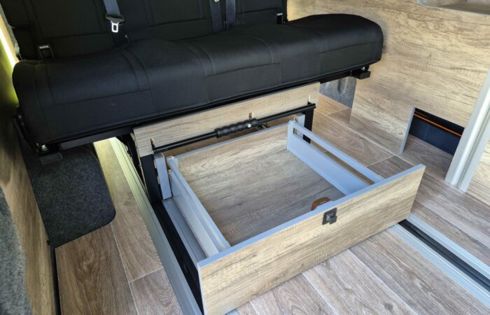 Un tiroir coulissant en bois est ouvert sous une banquette noire à l'intérieur d'un camping-car avec un plancher et des panneaux en bois.