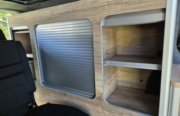 Étagère en bois intégrée à l'intérieur d'une camionnette, comprenant des étagères ouvertes à droite et un grand compartiment avec une porte roulante argentée fermée à gauche, à côté d'un siège noir.