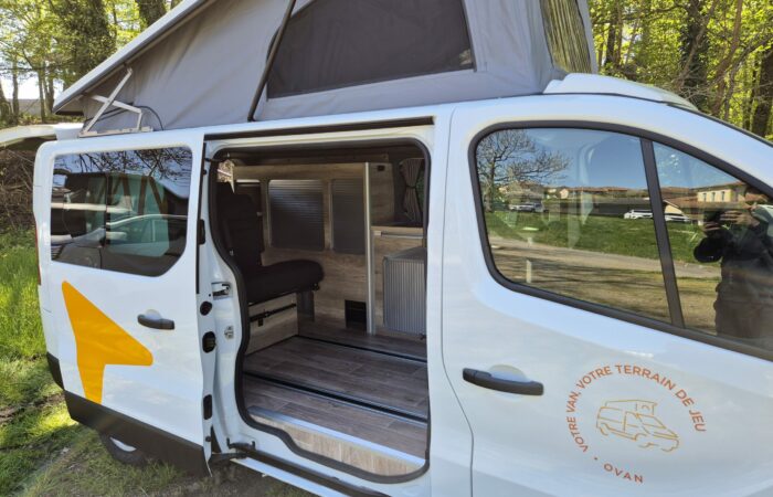 Un camping-car blanc dont la porte latérale est ouverte, révélant un intérieur compact avec banquette, rangements et boiseries. Le toit est surélevé pour une hauteur sous plafond accrue. Des arbres et de l'herbe sont visibles en arrière-plan.