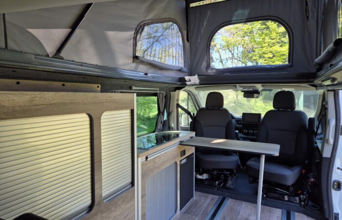 Intérieur de camping-car moderne avec toit relevable, deux sièges avant pivotants, une petite table et une kitchenette compacte. De grandes fenêtres laissent entrer la lumière du soleil, créant un espace lumineux et chaleureux, entouré de verdure.