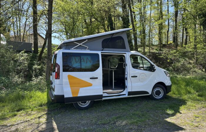 Un camping-car blanc à toit relevable est garé sur une pelouse en bordure d'une forêt. La porte latérale est ouverte, révélant l'intérieur. La lumière du soleil filtre à travers les arbres en arrière-plan.