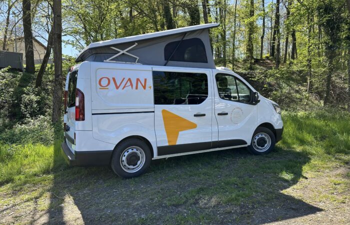 Un camping-car blanc avec la marque « OVAN » et une flèche jaune est garé sur l'herbe près d'une zone boisée, avec son toit relevable étendu sous la lumière du soleil.