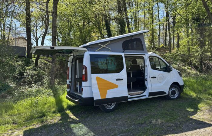 Un camping-car blanc avec ses portes arrière et latérales ouvertes et le toit relevé est garé sur une zone herbeuse entourée de grands arbres verts à la lumière du jour.