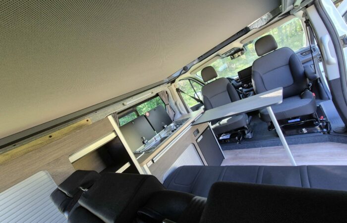 Intérieur d'un camping-car avec deux sièges avant, une petite table, une kitchenette avec évier et des placards. Le toit surélevé laisse entrer la lumière naturelle par les fenêtres.