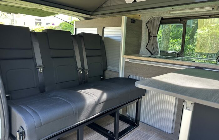 Intérieur d'un camping-car avec banquette noire et ceintures de sécurité, table pliante, plancher imitation bois et fenêtres avec rideaux. La lumière pénètre à flots, soulignant le design moderne et compact.