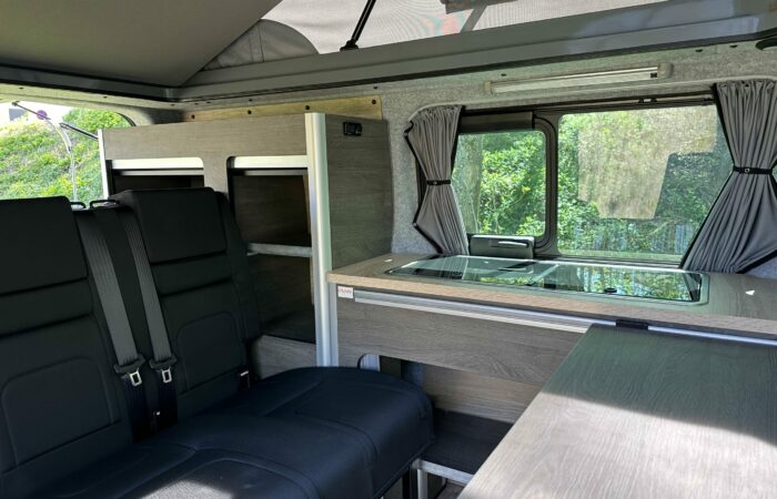 Intérieur d'un camping-car moderne comprenant des sièges noirs avec ceintures de sécurité, un coin cuisine compact avec un évier, des armoires en bois et une fenêtre avec des rideaux donnant sur le feuillage vert à l'extérieur.