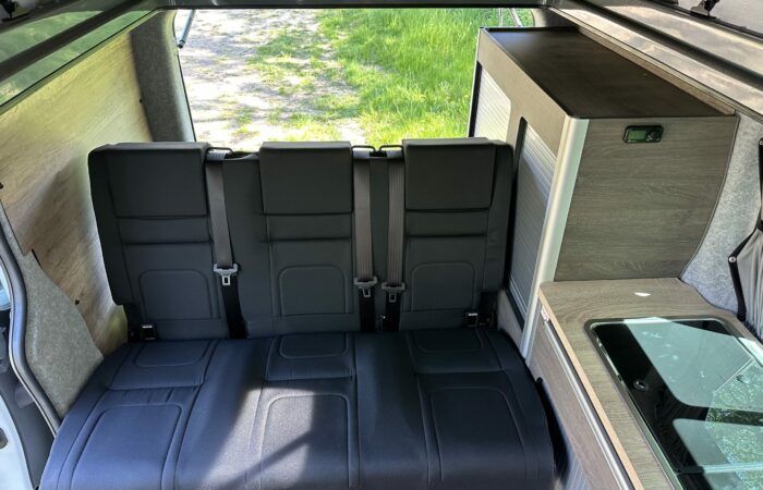 Intérieur d'un camping-car avec une banquette noire rabattue, des armoires en bois intégrées, un petit évier recouvert de verre et la lumière du soleil traversant la porte ouverte sur l'herbe verte à l'extérieur.