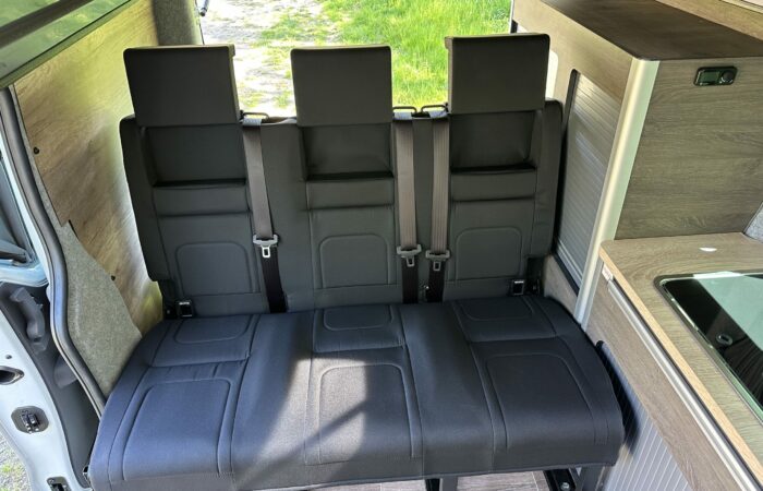 Trois sièges noirs rembourrés avec appuis-tête et ceintures de sécurité à l'intérieur d'un camping-car, à côté d'un petit coin kitchenette avec un évier et un comptoir ; de l'herbe verte est visible à l'extérieur de la porte ouverte.