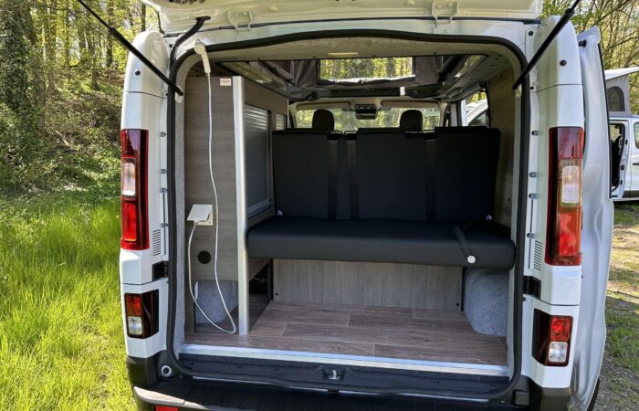 L'arrière d'un camping-car blanc avec ses portes arrière ouvertes, montrant une banquette noire, un parquet en bois et une étagère intégrée avec des câbles de chargement, garé sur l'herbe près d'une zone boisée.