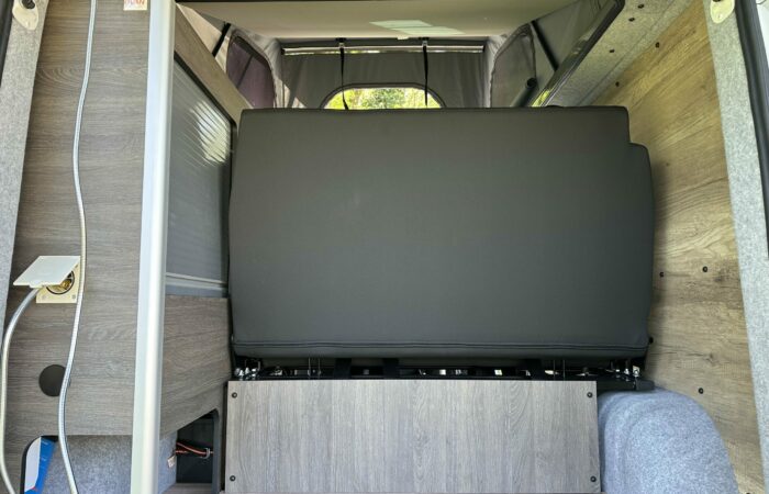 Intérieur d'un camping-car montrant l'arrière d'une banquette noire rabattue, des boiseries, une prise de courant et une fenêtre avec des arbres verts visibles à l'extérieur. Le sol est en bois et les parois sont recouvertes de tissu gris.